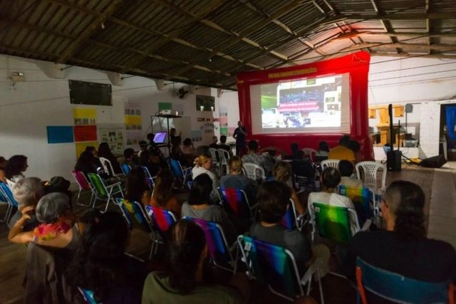 Salão comunitário de telhado metálico com paredes cobertas de cartazes; dezenas de pessoas em cadeiras de praia assistem a um filme numa tela inflável vermelha com a marca da produtora.