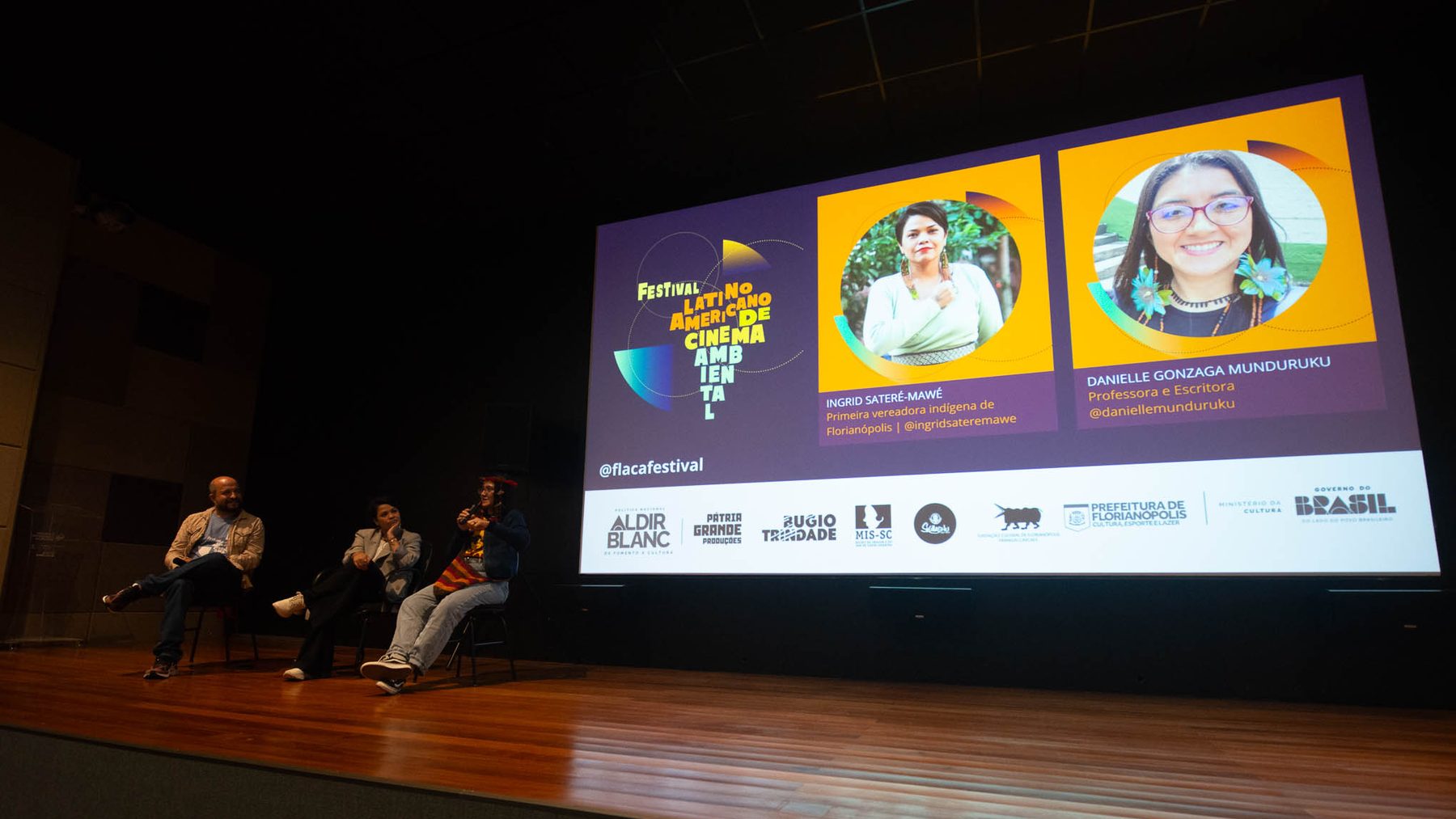 Três pessoas sentadas em roda de conversa sobre um palco de madeira, diante de uma tela que projeta a identidade do Festival Latino-Americano de Cinema Ambiental, os cards de duas convidadas e a faixa de apoiadores.