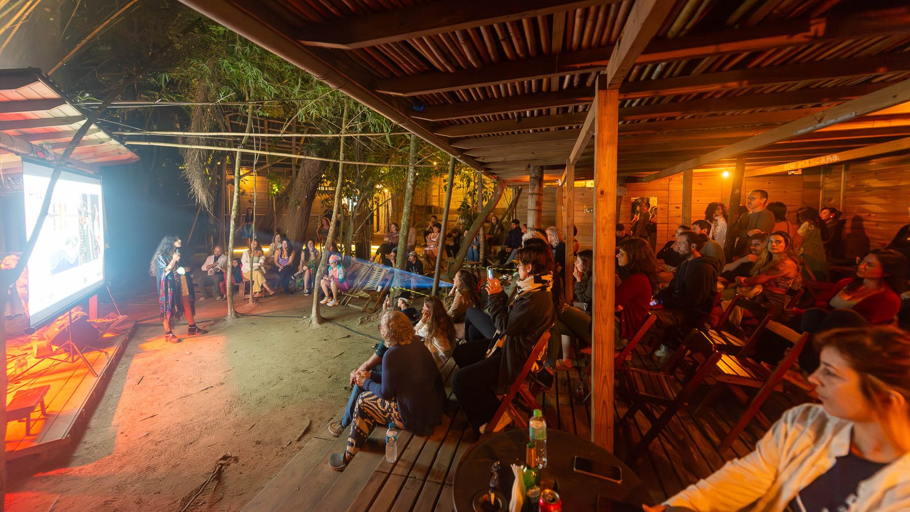 Sessão de cinema ao ar livre à noite: tela iluminada à esquerda, duas pessoas conduzem uma conversa ao microfone e o público acompanha sentado em cadeiras sobre areia e deque de madeira.